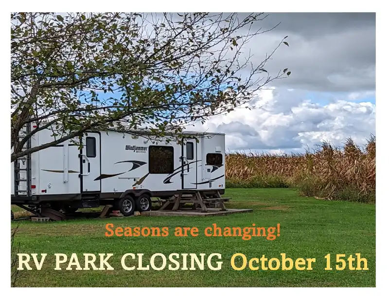 A recreational vehicle (RV) parked on grass next to a picnic table, with a caption reading "Seasons are changing! RV PARK CLOSING October 15th" against an autumnal backdrop of trees and a cornfield under a partly cloudy sky.