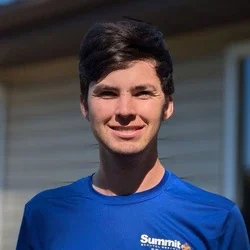 A young man with dark hair smiling and wearing a blue t-shirt standing outdoors.